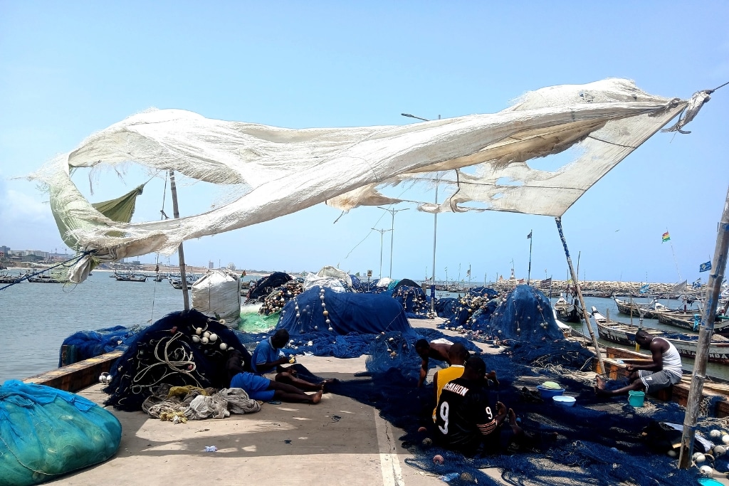 Fishermen sit in the patchy shade of a tattered tarp as the midday heat beats down.